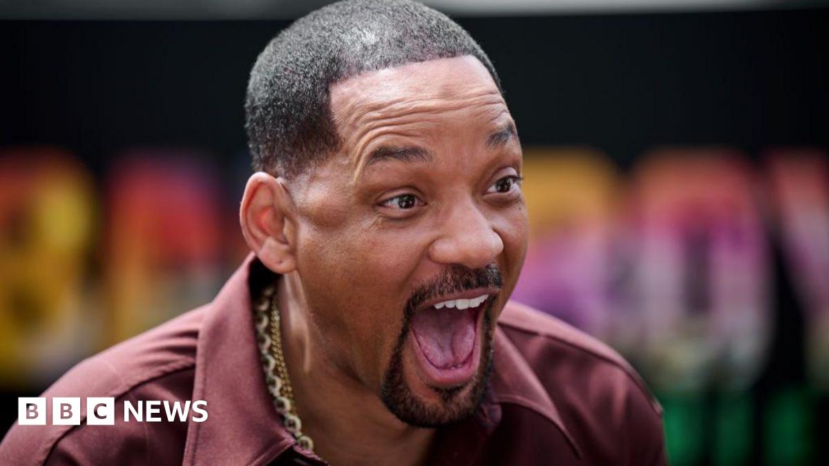 Will Smith is looking slightly to his right with an animated expression on his face: mouth wide open showing his tongue and upper teeth in a broad smile with his eyebrows high on his forehead. He has short cropped hair shaved into sharp edges, and is wearing an open necked burgundy shirt with broad lapels. He has several thick gold chains around his neck.